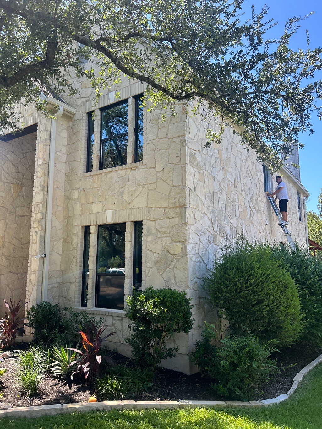 Glacier Home Exteriors installer on ladder installing Anlin windows with black frames on 2-story stone home in Lakeway TX action shot
